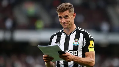 Mid-30s male Newcastle United staff member, athletic build, waist-up, wearing the black and white home shirt with the...