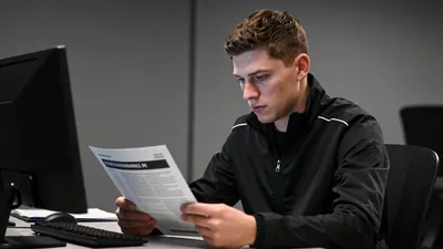 A mid-30s anonymous Newcastle United staff member, waist-up, reviewing scouting reports in a dimly lit office;