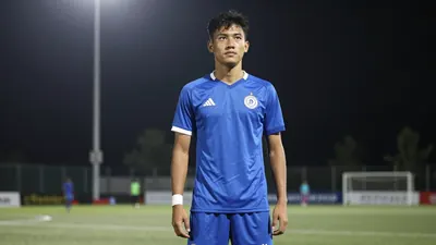 A 24-year-old midfielder in a solid blue shirt and shorts, standing on a football pitch with a determined expression