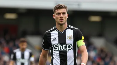 A determined 25-year-old Newcastle United left-back with a strong build, wearing the black and white home kit with the...