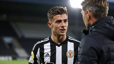 A young Newcastle United player in a black and white striped kit, mid-conversation with a coach in a training session