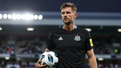 A photo of a 38-year-old German coach, head-and-shoulders, waist-up, with a football in hand, wearing a black jersey with...