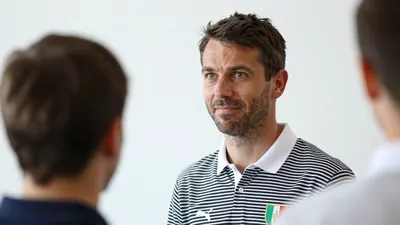 A 30-year-old German head coach, wearing a black and white striped shirt with a white collar, stands in front of a white...