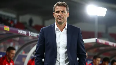 A 38-year-old German coach in a dark blue suit and white shirt, standing on the sidelines during a match, focused expression